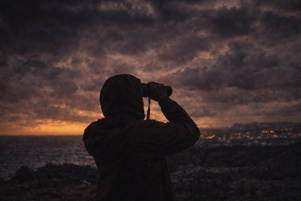 Figura solitária observando o horizonte com binóculos sob um céu escuro e tempestuoso, simbolizando o medo humano diante do desconhecido.